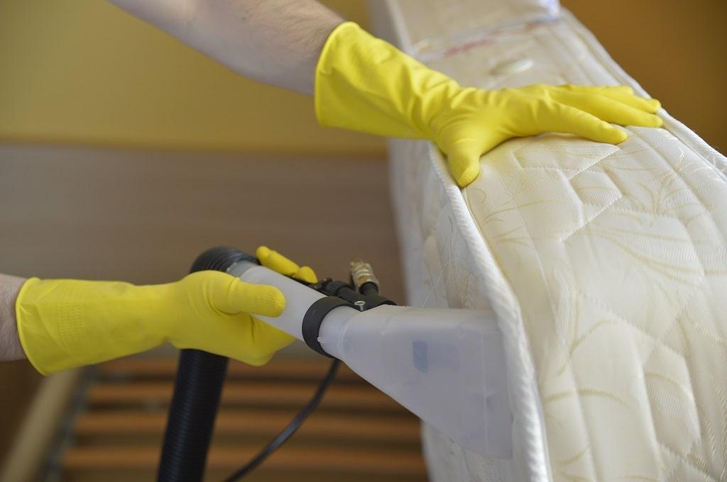 ndis mattress cleaner wearing yellow rubber gloves, holding a mattress up with one hand and using a machine to clean it with the other
